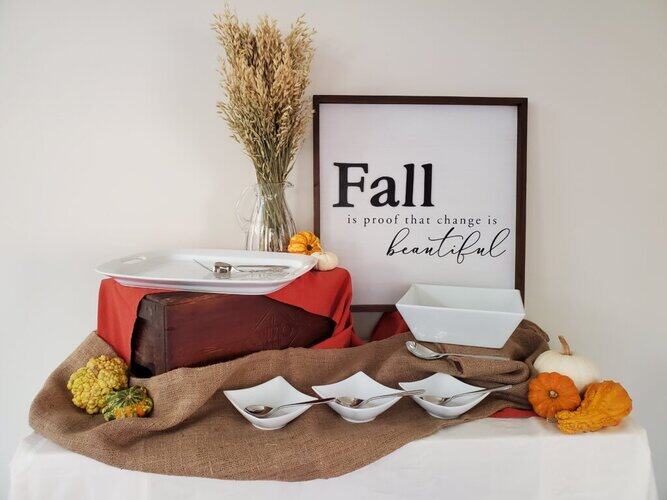 Vase with dried autumn grasses, fall-themed pumpkins, and a decorative fall quote frame on a white table decorated with warm-colored textiles.